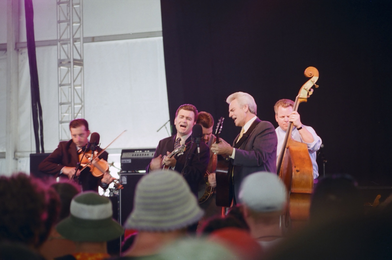 A suited bluegrass band with fiddle, mandolin, guitar and upright bass sings together at a microphone inside a festival tent, hats of the crowd in the foreground.